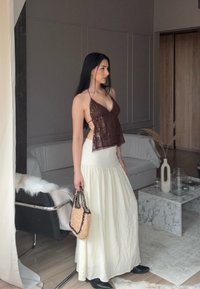Donna dai lunghi capelli scuri che indossa un top halter in paillettes color marrone, una gonna maxi crema, stivali neri e tiene in mano una borsa intrecciata in un soggiorno moderno.