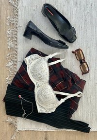 Hvid blonde-bh, ternet nederdel, sorte bukser med striber, to sorte hæle og brune mønstrede solbriller på et tekstureret hvidt tæppe.