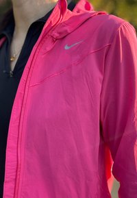 Kerge, ere roosa Nike jope kapuutsiga, millel on hõbedane logo ja kontrastne must sisekihte. Sile tekstuur, minimalistlik disain.