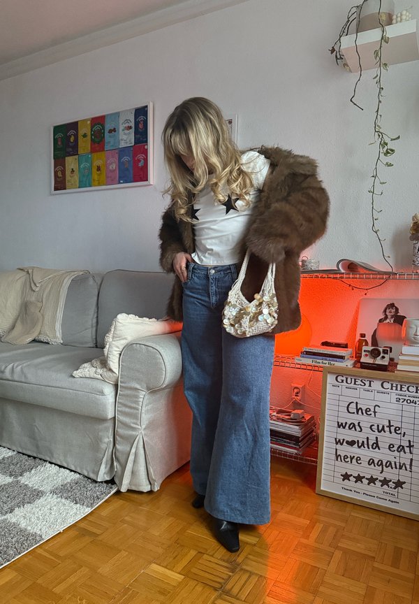 Vrouw met lang blond haar draagt een bruine bontjas, een wit topje met sterretjes, wijde jeans en houdt een met kralen versierde handtas vast in een gezellige woonkamer.