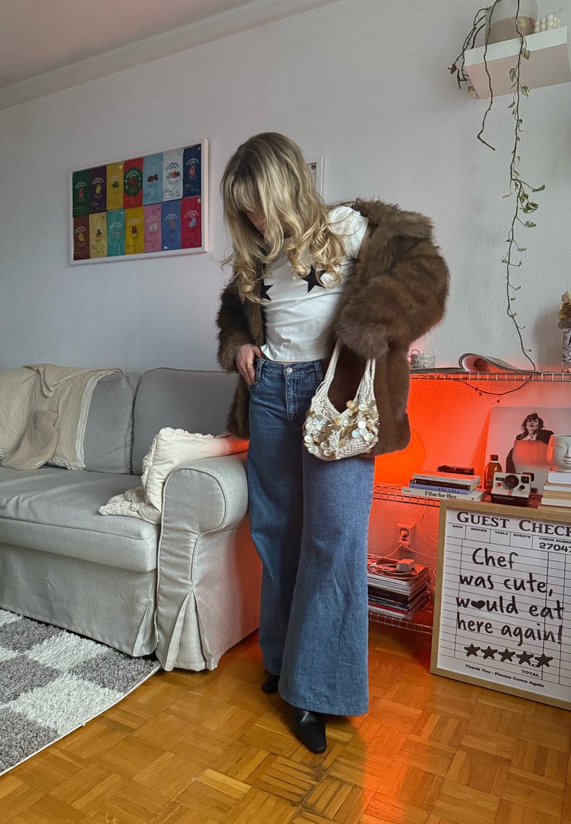 Femme aux longs cheveux blonds porte un manteau en fourrure marron, un haut blanc à motifs étoilés, un jean à jambes larges et tient un sac à main perlé dans un salon confortable.