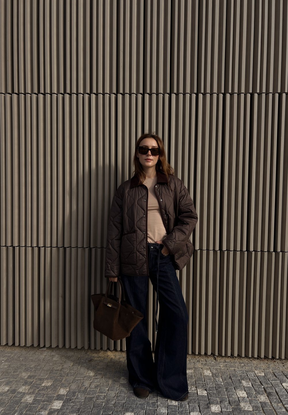 Brun quiltet jakke, beige topp, mørke vide jeans og håndveske. Solbriller og langt hår. Teksturerte bakgrunn av vertikale linjer.
