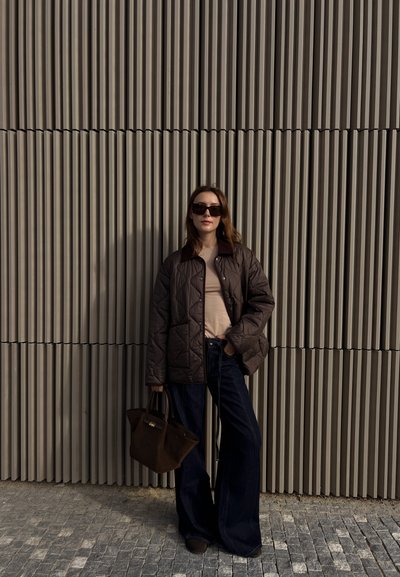 Chaqueta acolchada marrón, blusa beige, jeans de campana oscuros y bolso. Gafas de sol y cabello largo. Fondo texturizado de líneas verticales.