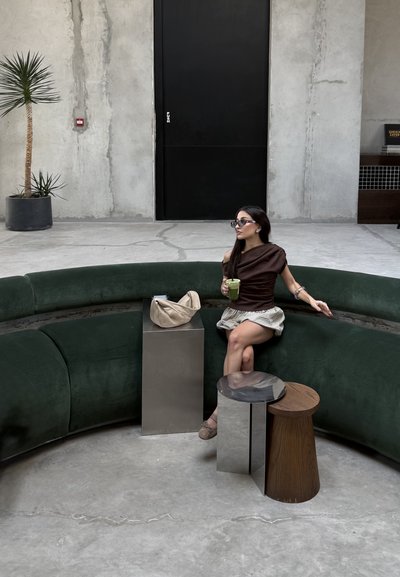Mujer con gafas de sol sentada en un sofá curvo verde oscuro sosteniendo una bebida verde en una habitación minimalista con paredes y suelo de hormigón.