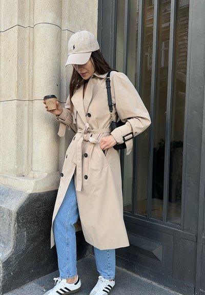 Trench beige con la cintura atada, botones negros, llevado sobre jeans azul claro y zapatillas en blanco y negro. Sosteniendo una taza de café.