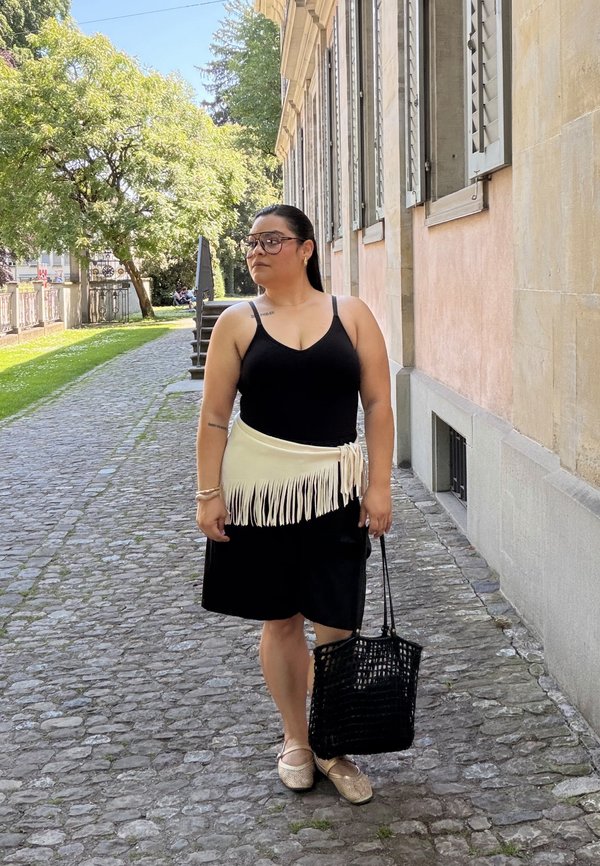 Žena v čiernych šatách a béžovom opasku s strapcami stojí na dláždenej ceste vedľa budovy s okenicami, drží čiernu pletenú tašku.