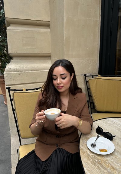 Blazer marrón sobre una falda negra plisada, sosteniendo una taza de café blanca. Cerca, un plato blanco con una cuchara y un caramelo de caramelo sobre una mesa de mármol.