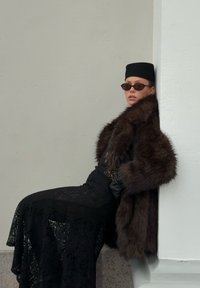 Brun pelsimitasjekk over svart blonderkjole, svarte hansker og solbriller. Modellen lener seg mot en lys grå vegg.