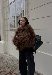 Donna con giacca marrone pelosa e paraorecchie neri, in piedi con le braccia incrociate accanto a un edificio beige con finestra.