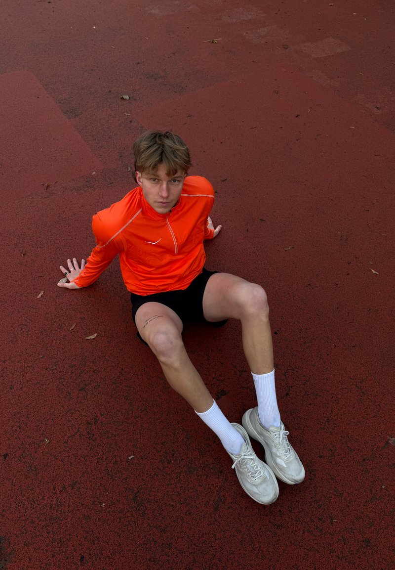 Leuchtend orangefarbenes langärmeliges Sportshirt mit reflektierenden Akzenten, schwarze Shorts, weiße Socken und Schuhe, sitzend auf einer roten Laufbahn.