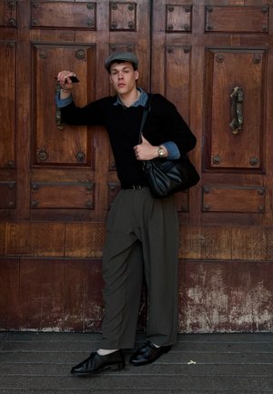 Joven con gorra gris, suéter negro, pantalones anchos de color oliva, zapatos negros y un bolso al hombro se apoya en una gran puerta de madera sosteniendo un teléfono.