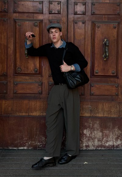 Joven con gorra gris, suéter negro, pantalones anchos de color oliva, zapatos negros y un bolso al hombro se apoya en una gran puerta de madera sosteniendo un teléfono.