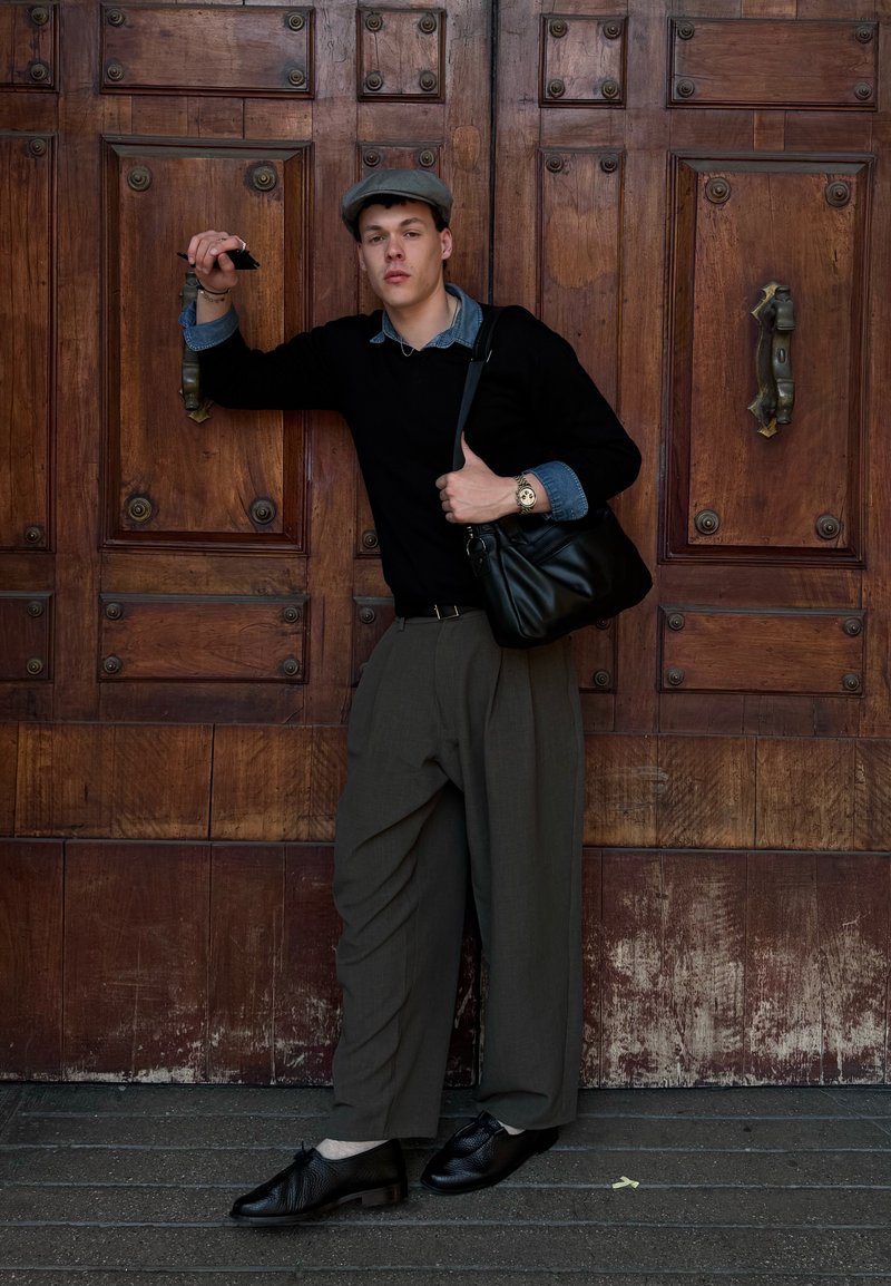 Νεαρός άνδρας με γκρι καπέλο, μαύρο πουλόβερ, φαρδιά ελιά παντελόνια, μαύρα παπούτσια και τσάντα ώμου στηρίζεται σε μια μεγάλη ξύλινη πόρτα κρατώντας ένα τηλέφωνο.