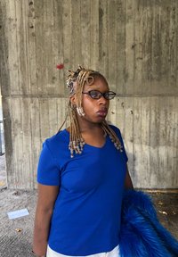 Das Modell trägt ein blaues Baumwoll-T-Shirt mit V-Ausschnitt, kombiniert mit einem blauen Kunstpelz-Accessoire und kunstvoll geflochtenem Haar, das mit Perlen geschmückt ist.