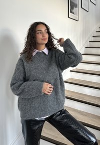 Grijze, pluizige trui met brede mouwen, over een lichtroze overhemd met kraag; glanzende zwarte leren broek met krokodillenpatroon.