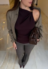 Haut sans manches bordeaux avec un design torsadé et des accessoires dorés, associé à un blazer à motif pied-de-poule, un sac en tissage marron et un pantalon gris ajusté.