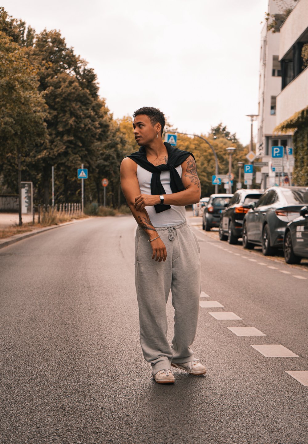 Άτομο που φοράει λευκό κοντομάνικο μπλουζάκι και γκρι παντελόνια φόρμας, με μια μαύρη ζακέτα δεμένη στους ώμους. Αστική σκηνή με παρκαρισμένα αυτοκίνητα.