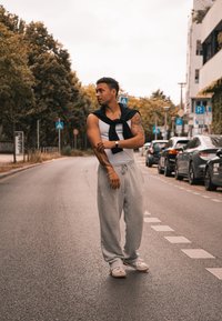 Άτομο που φοράει λευκό κοντομάνικο μπλουζάκι και γκρι παντελόνια φόρμας, με μια μαύρη ζακέτα δεμένη στους ώμους. Αστική σκηνή με παρκαρισμένα αυτοκίνητα.