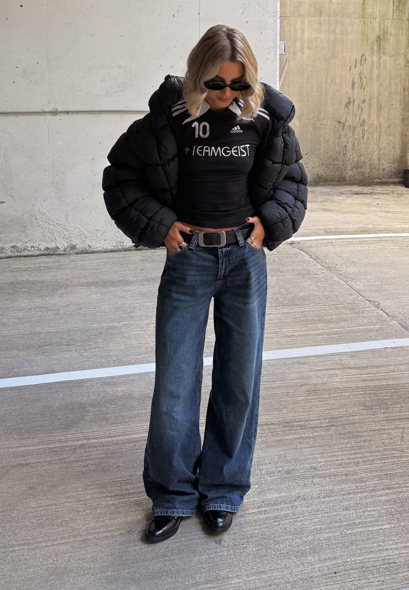 Černá péřová bunda, černé sportovní tričko s bílými akcenty, modré široké džíny a lesklé černé boty. Doplňky zahrnují sluneční brýle.