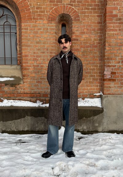 Hombre con bigote, vestido con abrigo largo, suéter oscuro, bufanda y vaqueros, de pie sobre la nieve frente a una pared de ladrillo rojo con ventanas arqueadas.