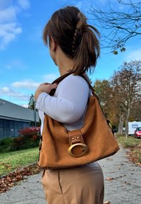 Braune Wildleder-Handtasche mit einem goldenen, kreisförmigen Akzent, einem Schulterriemen aus Stoff und einem gepassten Design, platziert vor einem Hintergrund aus blauem Himmel und Bäumen.