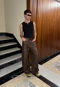 Man wearing black sleeveless buttoned shirt, brown wide-leg pants, yellow sneakers, and sunglasses leaning against wall by stairs.