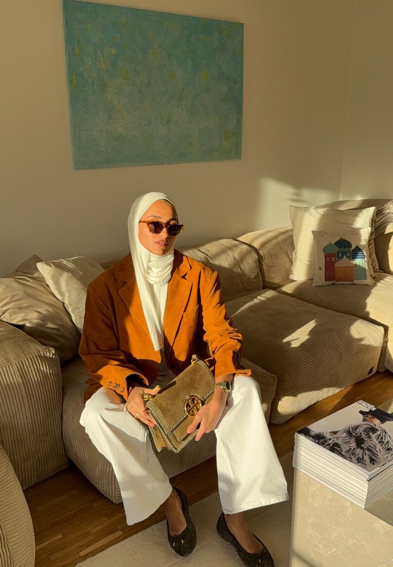 Mannequin portant un blazer en daim marron, un haut blanc et un pantalon blanc ample. Tenant un sac à main marron avec des coutures décoratives. En arrière-plan, un tableau bleu.