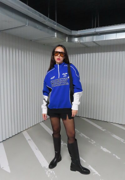 Sudadera azul con cierre y detalles blancos, con texto gráfico, combinada con una minifalda negra y botas negras hasta la rodilla. Incluye gafas de sol de gran tamaño.