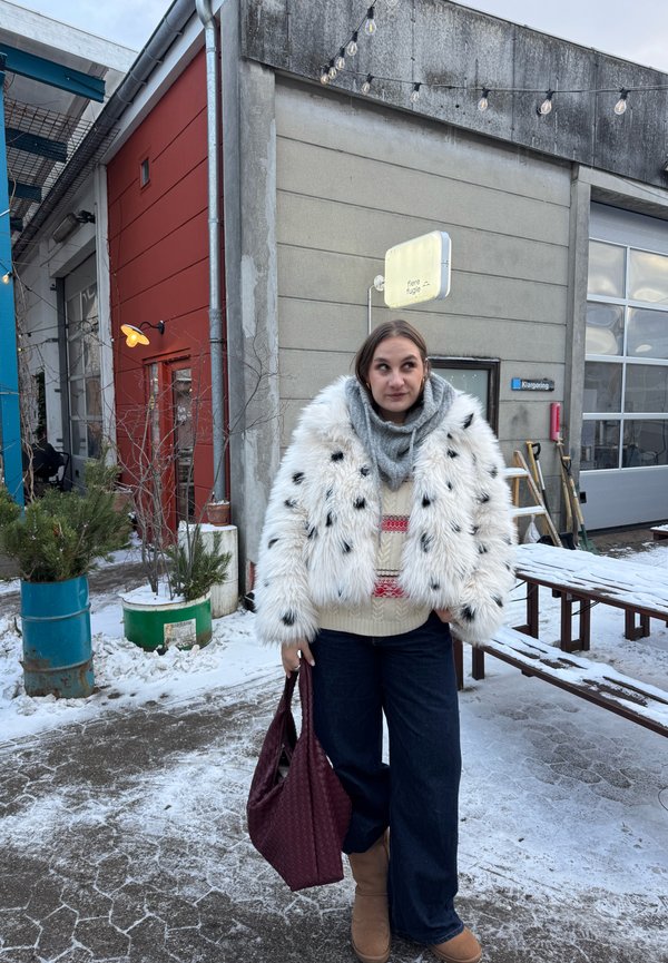 Žena u bijelom krznenom kaputu, šal i čizmama drži bordo torbu, stoji na snježnom tlu pokraj industrijskih zgrada s vanjskim svjetlucavim lampicama.