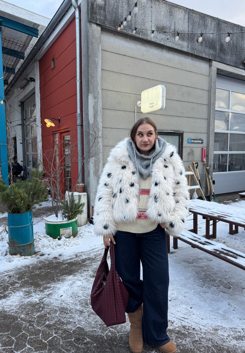 Femme en manteau de fourrure blanc, écharpe et bottes tenant un sac bordeaux, debout sur un sol enneigé près de bâtiments industriels avec des guirlandes lumineuses extérieures.