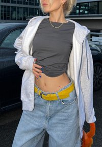 Camiseta gris corta debajo de una sudadera blanca con cremallera, combinada con jeans de tiro alto en azul y un cinturón amarillo brillante. Uñas largas con esmalte azul.
