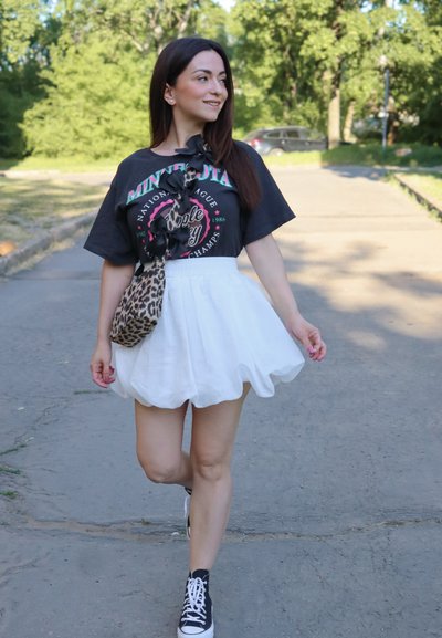 Camiseta gráfica negra con texto colorido, falda acampanada blanca, bolso cruzado de estampado animal y zapatillas altas negras en un sendero.