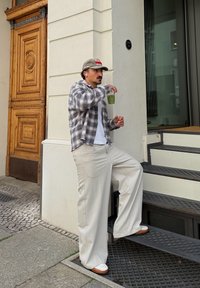 Pantalones beige oversize, una camisa de franela a cuadros en negro y gris, y una gorra gris. Sosteniendo una bebida verde, de pie junto a unos escalones con superficie texturizada.