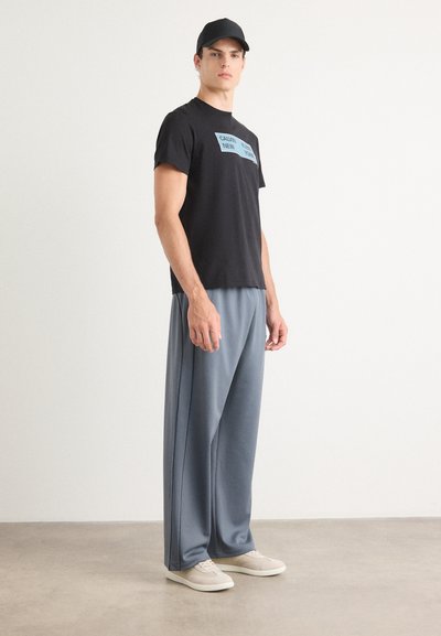 Camiseta de algodón negra con texto azul, combinada con pantalones holgados de color gris claro y zapatillas beige, exhibida sobre un fondo neutral.