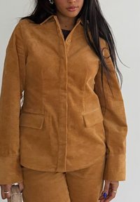 Femme portant une veste en velours côtelé marron avec des poches à rabat et un pantalon assorti, tenant un petit sac beige clair.