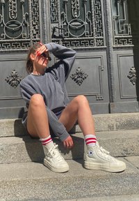 Sneakers brancos de cano alto com sola grossa, combinados com meias de tornozelo listradas em vermelho e branco. A pessoa veste um moletom cinza e está sentada em degraus de pedra.