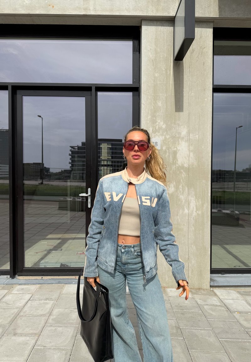 Femme aux longs cheveux blonds portant des lunettes de soleil rouges, une veste en denim claire, un crop top, un jean large, tenant un sac à main noir, debout à l'extérieur.