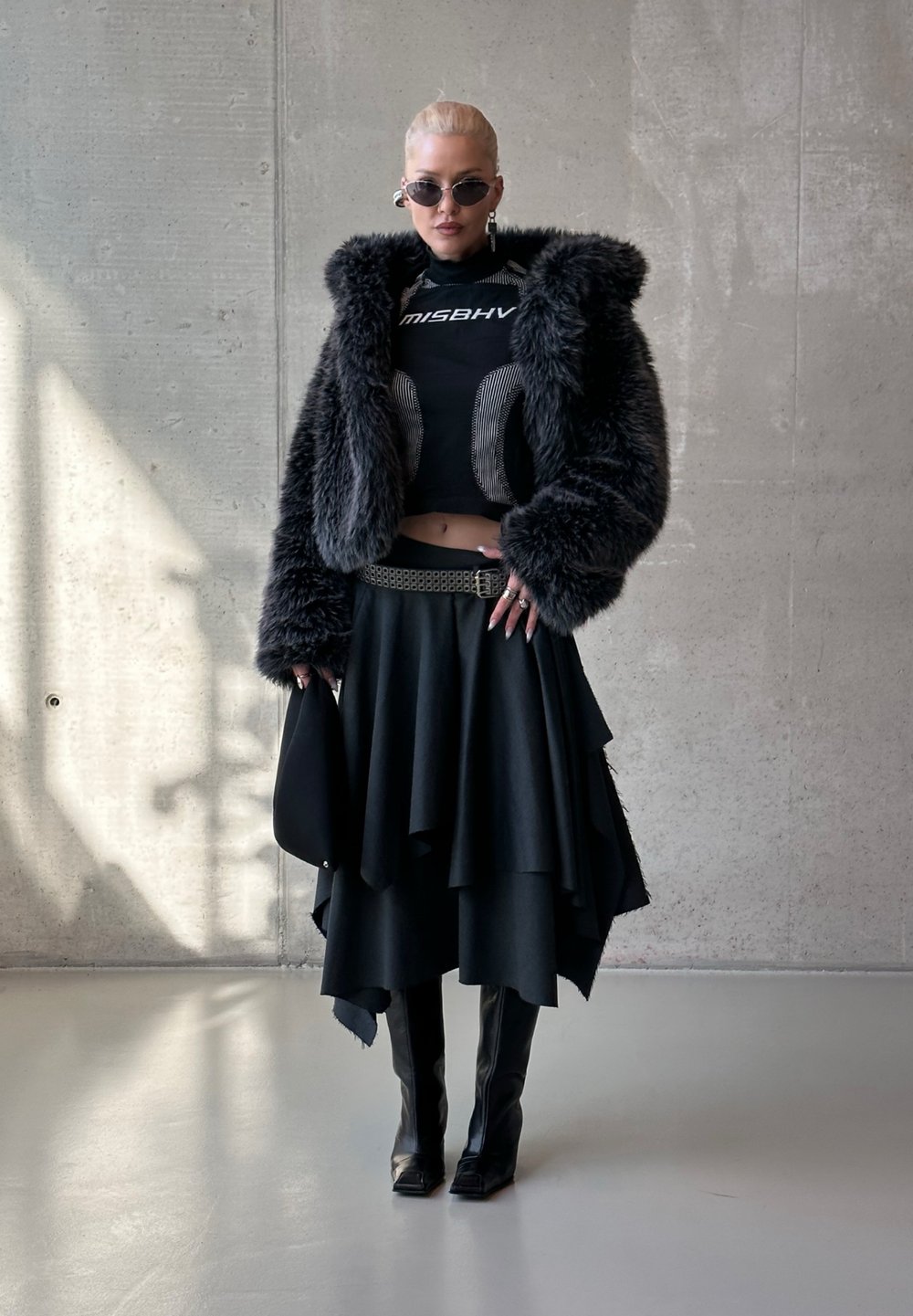 Personne portant une veste en fausse fourrure noire, un top court graphique, une jupe asymétrique à plusieurs niveaux, des bottes montant aux genoux, des lunettes de soleil, et tenant un sac à main noir.