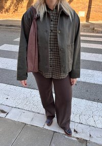 Giacca in tela verde, camicia a quadri marrone, pantaloni marrone scuro e scarpe marroni. Accessorio: borsa a tracolla bordeaux. Sfondo di una strada chiara.