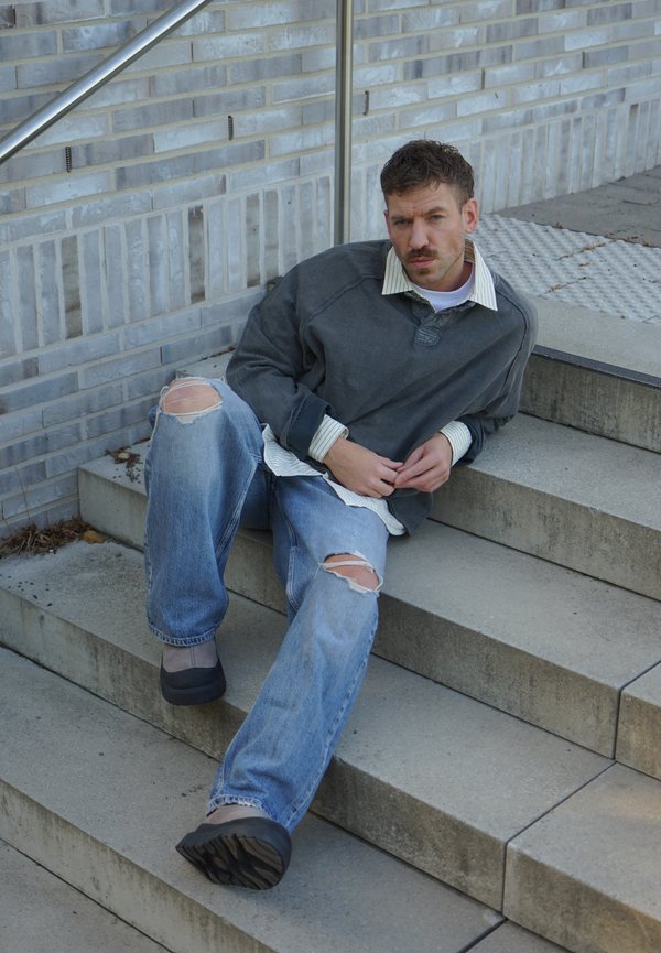 Un homme moustachu, vêtu d’un pull gris et d’un jean déchiré, est assis sur des marches en pierre à l’extérieur, à côté d’un mur de briques.