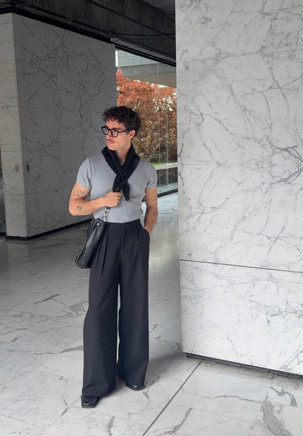 Un homme portant des lunettes et un sac en bandoulière se tient dans un hall en marbre. Il porte une chemise grise et un pantalon noir ample.