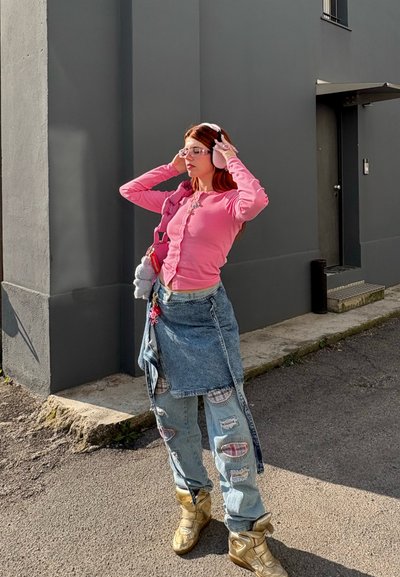 Top de manga larga rosa, falda de denin con capas, jeans ligeros rasgados de color azul con parches y botas metálicas doradas. Los accesorios incluyen orejeras y gafas.