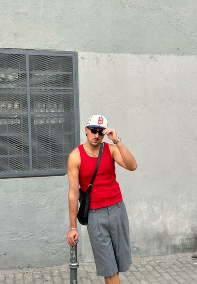Camiseta sin mangas roja con textura acanalada, shorts grises plisados, bolso cruzado negro, sombrero blanco con logo rojo, gafas de sol, apoyada contra un poste. Fondo de pared con textura.