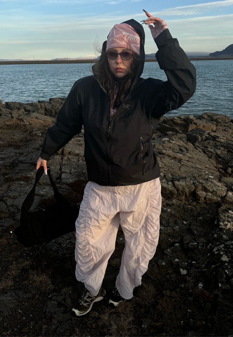Person, die eine schwarze Kapuzenjacke, ein pink gemustertes Kopftuch, eine Sonnenbrille und weiße, geraffte Hosen trägt, steht am felsigen Ufer mit einer schwarzen Tasche in der Hand nahe am Wasser.