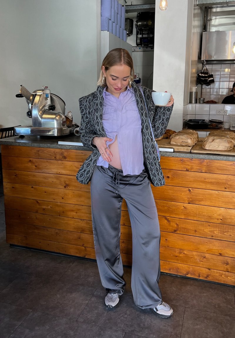 Ανοιχτό μωβ πουκάμισο κάτω από ένα καλυμμένο μαύρο και άσπρο σακάκι, συνδυασμένο με γκρι σατέν παντελόνι και αθλητικά παπούτσια, κρατώντας έναν καφέ στο χέρι.