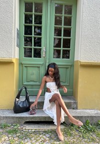 Abito bianco in pizzo con dettaglio a taglio, borsa hobo nera e ballerine color nud. Seduta su gradini di pietra di fronte a porte verdi a doppio battente.