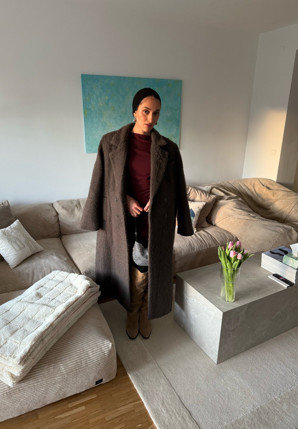 Femme en manteau foncé et foulard sur la tête se tient dans un salon avec un canapé beige, des couvertures pliées, une table basse en marbre et un vase de tulipes roses.