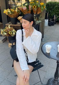 Λευκή υφασμάτινη μπλούζα με μακριά μανίκια, γιακά και λεπτομέρεια με φιόγκο, σε συνδυασμό με μαύρες βερμούδες. Ρυθμός καφέ σε εξωτερικό χώρο με ανθοδέσεις.