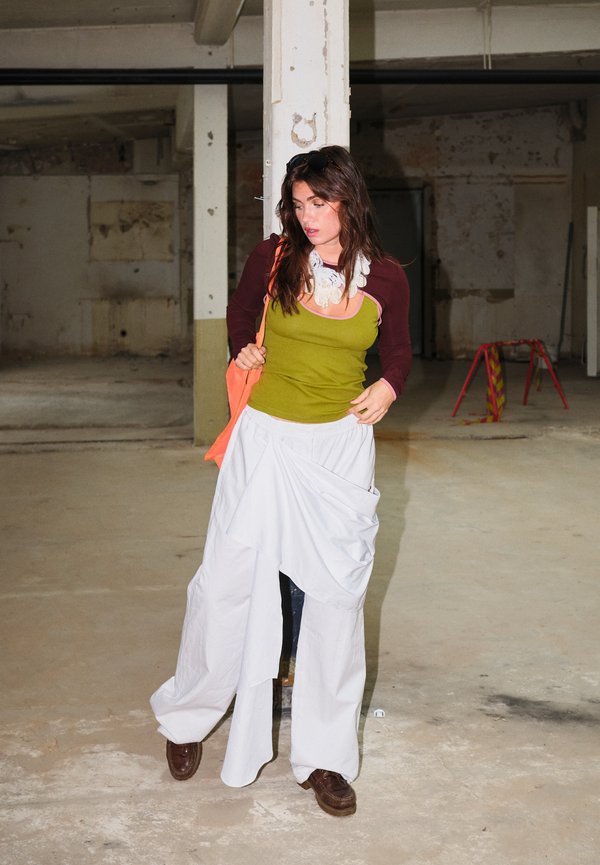 Una mujer con una prenda superior verde y unos pantalones blancos vaporosos camina por un espacio industrial vacío con un bolso naranja al hombro.