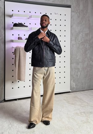 Hombre ajustándose una chaqueta de cuero negra, vistiendo pantalones anchos beige y zapatos negros, de pie cerca de un panel de clavijas blanco con ropa y zapatos exhibidos.